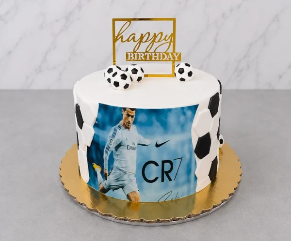 Pastel redondo con cubierta blanca y detalles de hexágonos de fondant formando un patrón de balón de fútbol en los costados. En el centro presenta una gran imagen comestible de Cristiano Ronaldo (CR7) con el uniforme del Real Madrid. Está coronado con tres pequeños balones de fútbol y un letrero geométrico dorado de "happy BIRTHDAY". Castillo's Cake Shop.
