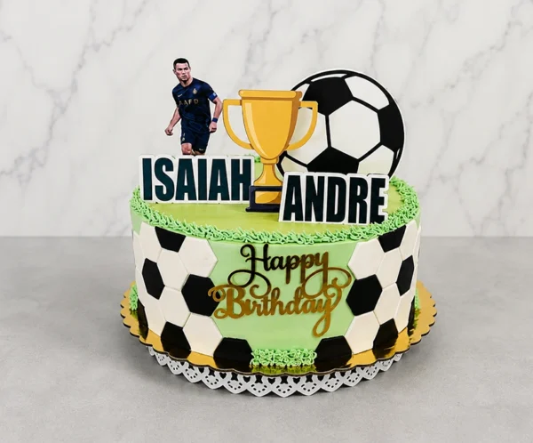 Pastel con cubierta verde lisa y bordes de glaseado que simulan césped. Los laterales muestran un patrón parcial de balón de fútbol con hexágonos blancos y negros. Decorado al frente con un letrero dorado caligráfico de "Happy Birthday", y en la cima con recortes de los nombres "ISAIAH" y "ANDRE", un gran balón, un trofeo y la figura de Cristiano Ronaldo. Castillo's Cake Shop.