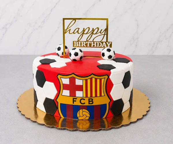 Pastel redondo con cubierta roja vibrante, adornado en los costados con hexágonos blancos y negros de fondant formando la clásica textura de un balón. Al frente destaca un gran escudo del FC Barcelona. En la parte superior lo decoran tres pequeños balones de fútbol y un letrero cuadrado dorado que dice "happy BIRTHDAY". Castillo's Cake Shop.