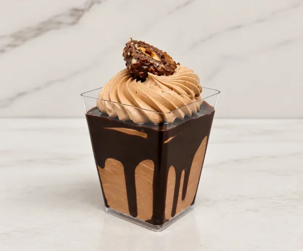 Ferrero en vasito. Elegante postre servido en un vasito cuadrado transparente. Se compone de una base de mousse o crema de chocolate con llamativos escurrimientos de jarabe de chocolate oscuro en el interior del vaso. La superficie está decorada con un gran rosetón de crema de chocolate claro y la mitad de un clásico chocolate Ferrero Rocher con avellanas.