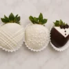 Tres jugosas fresas cubiertas de chocolate decoradas con temática de boda, presentadas individualmente en capacillos de papel blanco. La primera fresa tiene cobertura blanca con un elegante diseño de líneas cruzadas. La del centro es de color blanco espolvoreada con chispas de perlitas. La tercera fresa luce un detallado diseño de esmoquin en chocolate oscuro y blanco, completado con botones y un moño de chocolate.