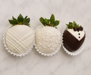 Tres jugosas fresas cubiertas de chocolate decoradas con temática de boda, presentadas individualmente en capacillos de papel blanco. La primera fresa tiene cobertura blanca con un elegante diseño de líneas cruzadas. La del centro es de color blanco espolvoreada con chispas de perlitas. La tercera fresa luce un detallado diseño de esmoquin en chocolate oscuro y blanco, completado con botones y un moño de chocolate.