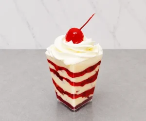 Fresas con crema en vasito. Postre servido en un vasito transparente cuadrado, compuesto por llamativas capas alternadas de crema blanca y mermelada o compota de fresa roja. Está coronado con un copo de crema batida y una clásica cereza roja brillante con tallo.