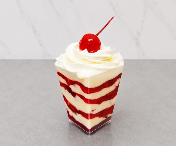 Fresas con crema en vasito. Postre servido en un vasito transparente cuadrado, compuesto por llamativas capas alternadas de crema blanca y mermelada o compota de fresa roja. Está coronado con un copo de crema batida y una clásica cereza roja brillante con tallo.