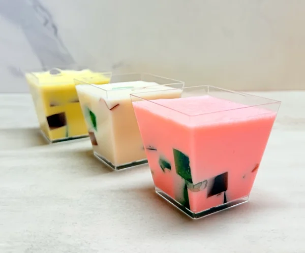 Gelatina 3 leches en vasito. Tres vasitos transparentes cuadrados que contienen gelatina de tres leches estilo mosaico. El primer vaso tiene una base de gelatina color rosa, el del medio es de color blanco y el del fondo es amarillo pálido. En su interior flotan coloridos cubos de gelatina de agua (verde, rojo y oscuro).