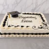 Elegante pastel rectangular tipo plancha con glaseado blanco. Presenta bordes clásicos de concha en color blanco y negro, decorado con arabescos negros, espolvoreado de perlas doradas comestibles, un birrete de graduación negro con borla dorada en una esquina y el mensaje "Congratulations Karina" en caligrafía negra. Castillo's Cake Shop.