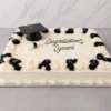 Pastel rectangular tipo plancha con cobertura blanca. Destaca por sus bordes superiores adornados con tupidos rosetones intercalados en blanco y negro, acompañados de sutiles destellos plateados. En la superficie incluye un birrete negro, un diploma enrollado y el mensaje central "Congratulations Dymari" en caligrafía negra. Castillo's Cake Shop.