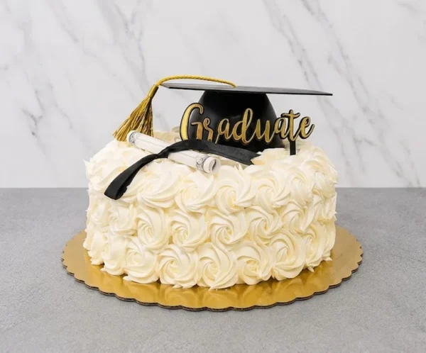 Pastel redondo de un piso completamente cubierto de hermosos rosetones de glaseado blanco (estilo rosette cake). En la cima está decorado de manera sobria con un birrete negro de graduación con borla dorada, un diploma enrollado con un listón negro y un elegante letrero dorado en acrílico que dice "Graduate". Castillo's Cake Shop.