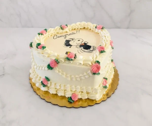 Hermoso pastel en forma de corazón con glaseado blanco y decoración vintage estilo Lambeth. Presenta bordes acanalados, guirnaldas y delicadas rosas rosadas de glaseado con hojas verdes. En el centro luce una tierna imagen comestible de Snoopy graduado saltando de felicidad, acompañada de la palabra "Congrats!!". Castillo's Cake Shop.