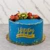 Pastel redondo cubierto con un vibrante glaseado azul, decorado con salpicaduras doradas y un borde superior irregular con detalles en pan de oro. En la corona presenta un fresco arreglo de frutas que incluye kiwi en forma de flor, fresas, uvas rojas y arándanos. Al frente luce un letrero amarillo que dice "Happy BIRTHDAY". Castillo's Cake Shop.