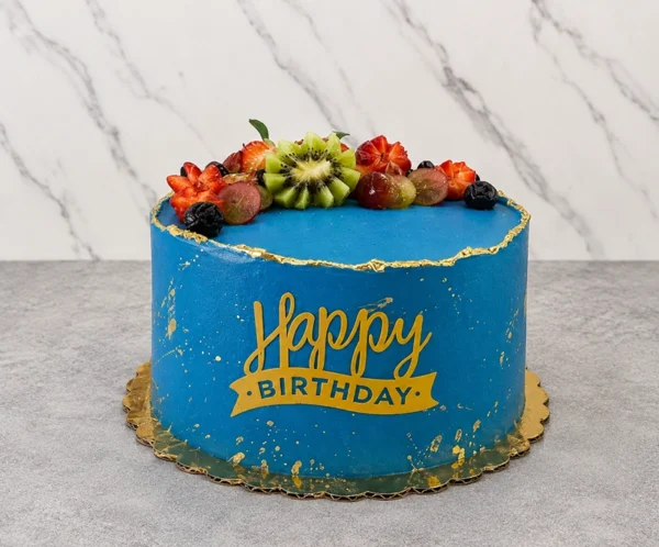 Pastel redondo cubierto con un vibrante glaseado azul, decorado con salpicaduras doradas y un borde superior irregular con detalles en pan de oro. En la corona presenta un fresco arreglo de frutas que incluye kiwi en forma de flor, fresas, uvas rojas y arándanos. Al frente luce un letrero amarillo que dice "Happy BIRTHDAY". Castillo's Cake Shop.