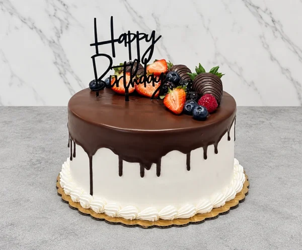 Delicioso pastel redondo con cobertura blanca lisa y un apetitoso escurrimiento (drip) de chocolate oscuro. La base está decorada con un borde clásico de concha en glaseado blanco. En la parte superior está adornado con fresas frescas, fresas cubiertas de chocolate, moras, arándanos, frambuesas y un elegante letrero acrílico negro de "Happy Birthday". Castillo's Cake Shop.