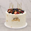 Elegante pastel redondo con cobertura blanca lisa, embellecido con delicadas perlas doradas y toques de hoja de oro esparcidos por los costados. La corona rebosa de una abundante mezcla de frutos rojos frescos, incluyendo fresas, zarzamoras y arándanos, acompañados de cinco velas doradas onduladas. Al frente destaca un letrero dorado en acrílico de "HAPPY Birthday". Castillo's Cake Shop.