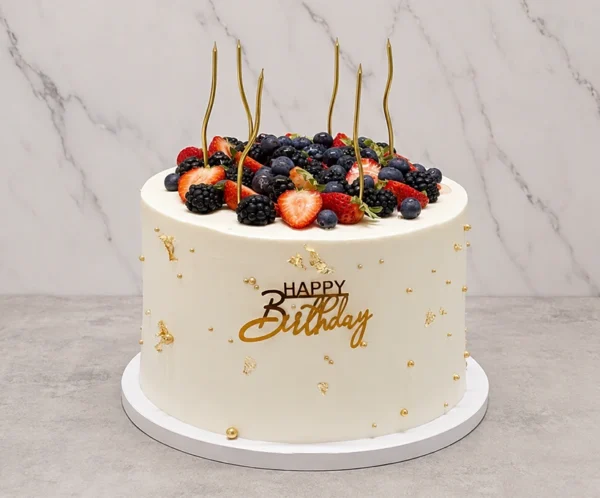 Elegante pastel redondo con cobertura blanca lisa, embellecido con delicadas perlas doradas y toques de hoja de oro esparcidos por los costados. La corona rebosa de una abundante mezcla de frutos rojos frescos, incluyendo fresas, zarzamoras y arándanos, acompañados de cinco velas doradas onduladas. Al frente destaca un letrero dorado en acrílico de "HAPPY Birthday". Castillo's Cake Shop.