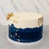 Moderno y sofisticado pastel redondo de un piso con un diseño bicolor. La mitad superior es de color blanco crema y la inferior azul marino profundo, divididas por una línea texturizada asimétrica tipo "falla". Está acentuado con detalles en hoja de oro, un elegante goteo (drip) dorado en un lateral y un racimo de esferas doradas en la parte superior izquierda. Castillo's Cake Shop.
