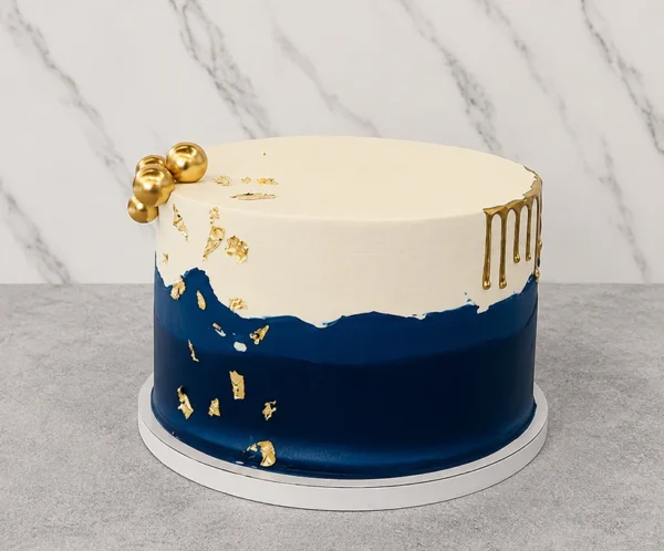 Moderno y sofisticado pastel redondo de un piso con un diseño bicolor. La mitad superior es de color blanco crema y la inferior azul marino profundo, divididas por una línea texturizada asimétrica tipo "falla". Está acentuado con detalles en hoja de oro, un elegante goteo (drip) dorado en un lateral y un racimo de esferas doradas en la parte superior izquierda. Castillo's Cake Shop.