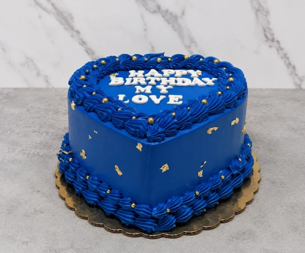 Romántico pastel en forma de corazón cubierto con un intenso glaseado azul rey. Está decorado con elaborados bordes entrelazados en la base y la corona, salpicado con pequeñas perlas doradas y delicados destellos de hoja de oro. En la superficie lleva el mensaje "HAPPY BIRTHDAY MY LOVE" en letras blancas gruesas. Castillo's Cake Shop.