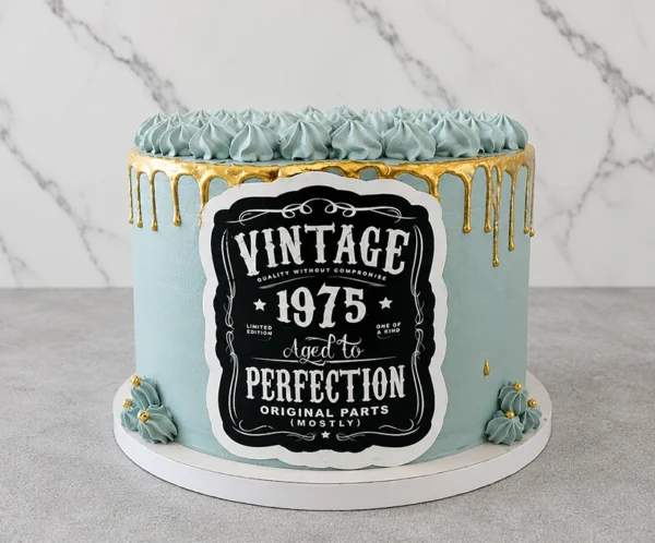 Pastel redondo con un elegante glaseado en tono azul grisáceo y un llamativo escurrimiento (drip) dorado. La corona está decorada con picos de glaseado del mismo tono azul. En el frente destaca una gran placa estilo etiqueta de whiskey vintage en negro y blanco con la frase "VINTAGE 1975 AGED TO PERFECTION ORIGINAL PARTS (MOSTLY)". Castillo's Cake Shop.