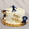 Pastel redondo con temática de fútbol en color blanco, adornado con bordes de concha, perlas y hoja de oro. En un costado luce un llamativo racimo de esferas en negro, blanco y dorado. Al frente lleva un letrero dorado de "Happy Birthday" y un recorte de camiseta azul marino con el nombre "BRIHAN" y el número "33". En la cima destaca la silueta de un jugador pateando un balón. Castillo's Cake Shop.