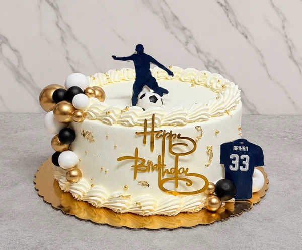 Pastel redondo con temática de fútbol en color blanco, adornado con bordes de concha, perlas y hoja de oro. En un costado luce un llamativo racimo de esferas en negro, blanco y dorado. Al frente lleva un letrero dorado de "Happy Birthday" y un recorte de camiseta azul marino con el nombre "BRIHAN" y el número "33". En la cima destaca la silueta de un jugador pateando un balón. Castillo's Cake Shop.