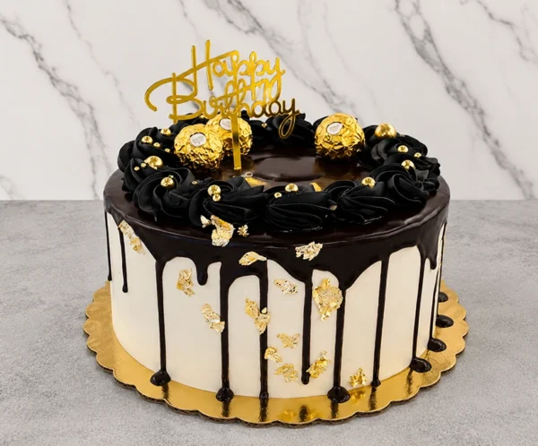 Elegante pastel redondo con cobertura blanca y un apetitoso escurrimiento (drip) de chocolate oscuro. Los costados están adornados con toques de hoja de oro. La corona está decorada con un anillo de tupidos rosetones de glaseado negro con perlas doradas, tres chocolates Ferrero Rocher y un letrero dorado superior de "Happy Birthday". Castillo's Cake Shop.