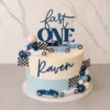 Elegante pastel de un piso con temática de carreras vintage. Presenta un diseño bicolor: la mitad superior es blanca y la inferior es azul claro con textura de líneas. En el centro resalta el nombre "Raven" en letras azul oscuro. Está adornado con autos de carreras retro de colores azul y blanco con el número "1", banderas a cuadros, racimos de esferas de chocolate simulando globos y un letrero superior que dice "fast ONE". Castillo's Cake Shop.