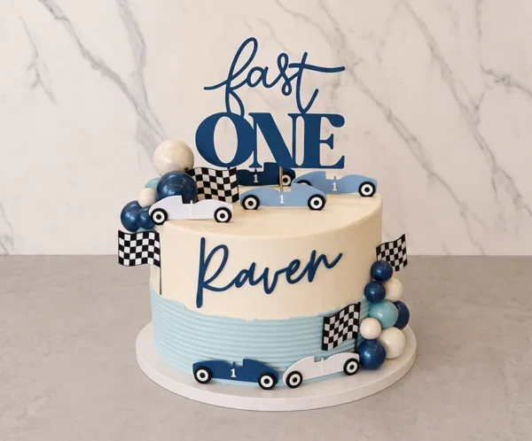 Elegante pastel de un piso con temática de carreras vintage. Presenta un diseño bicolor: la mitad superior es blanca y la inferior es azul claro con textura de líneas. En el centro resalta el nombre "Raven" en letras azul oscuro. Está adornado con autos de carreras retro de colores azul y blanco con el número "1", banderas a cuadros, racimos de esferas de chocolate simulando globos y un letrero superior que dice "fast ONE". Castillo's Cake Shop.