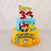 Pastel de dos pisos con temática de Pokémon. El piso inferior es de color amarillo con un delicado borde de perlas naranjas, decorado con el logotipo de Pokémon y las figuras de Bulbasaur, Squirtle y Charmander. El piso superior es azul claro y muestra a Ash Ketchum junto a Pikachu. En la corona destaca un adorno con el nombre "Henry", un número "3" rojo y una Pokebola. Castillo's Cake Shop.