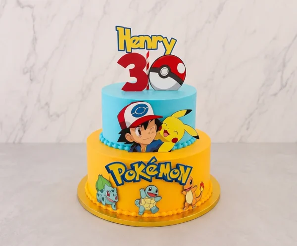 Pastel de dos pisos con temática de Pokémon. El piso inferior es de color amarillo con un delicado borde de perlas naranjas, decorado con el logotipo de Pokémon y las figuras de Bulbasaur, Squirtle y Charmander. El piso superior es azul claro y muestra a Ash Ketchum junto a Pikachu. En la corona destaca un adorno con el nombre "Henry", un número "3" rojo y una Pokebola. Castillo's Cake Shop.