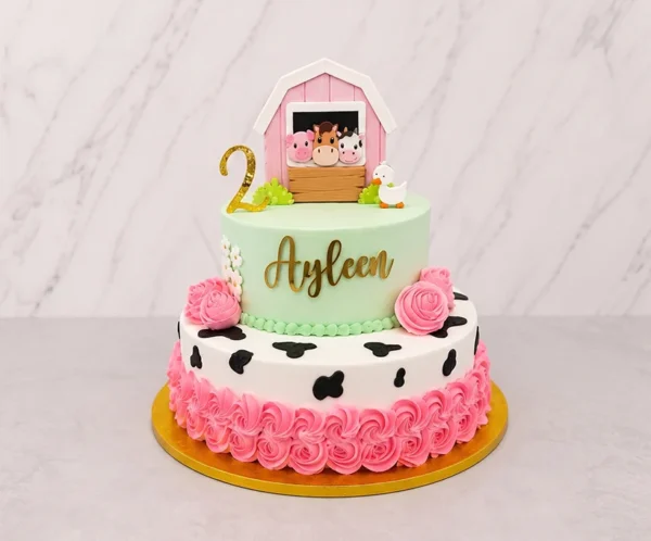 Tierno pastel de dos pisos con temática de animales de granja. El piso inferior es blanco con manchas de vaca negras y un abundante borde de rosetones en color rosa fuerte. El piso superior es verde menta pastel, lleva el nombre "Ayleen" en dorado y pequeñas margaritas. Está coronado con el número "2" dorado, un patito y una adorable figura de un granerito rosa asomando un cerdito, un caballo y una vaca. Castillo's Cake Shop.