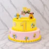 Pastel de dos pisos con dulce temática de abejitas. El piso inferior es rosa pastel y el superior es amarillo vibrante, ambos con textura de finas líneas horizontales y bordes de glaseado amarillo. Está decorado con figuras de simpáticas abejitas y el número "1" al centro. En la parte superior luce un adorno de panal, una ramita con flores rosadas, una abejita y el nombre "Mia". Castillo's Cake Shop.