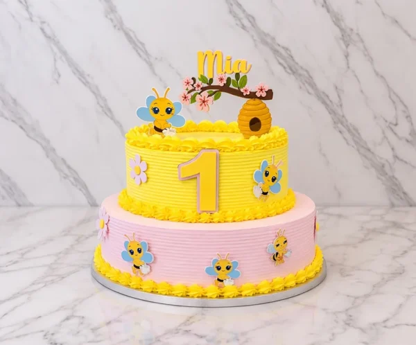 Pastel de dos pisos con dulce temática de abejitas. El piso inferior es rosa pastel y el superior es amarillo vibrante, ambos con textura de finas líneas horizontales y bordes de glaseado amarillo. Está decorado con figuras de simpáticas abejitas y el número "1" al centro. En la parte superior luce un adorno de panal, una ramita con flores rosadas, una abejita y el nombre "Mia". Castillo's Cake Shop.