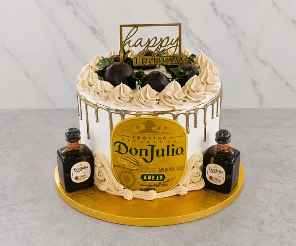 Elegante pastel redondo con cobertura crema, adornado con un espectacular escurrimiento (drip) dorado. En el centro frontal luce una gran etiqueta comestible de Tequila Don Julio Añejo. La corona está decorada con rosetones de crema, perlas doradas y fresas cubiertas de chocolate oscuro, rematada con un letrero dorado de "happy BIRTHDAY". En la base, a cada lado, reposan dos botellas miniatura reales de Tequila Don Julio. Castillo's Cake Shop.