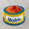 Atractivo pastel redondo envuelto con una oblea comestible que muestra el icónico logo de Cerveza Modelo Especial. Los bordes superior e inferior están decorados con rosetones entrelazados en colores azul y amarillo vibrante. La superficie superior está completamente cubierta con rodajas de kiwi fresco y fresas dispuestas en forma de flor, con una fresa entera en el centro. Castillo's Cake Shop.