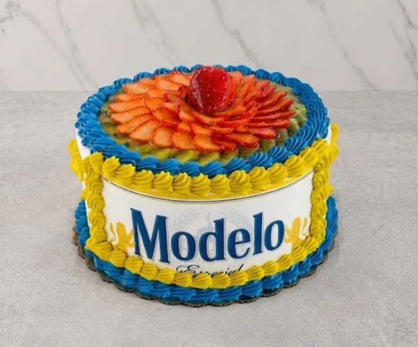 Atractivo pastel redondo envuelto con una oblea comestible que muestra el icónico logo de Cerveza Modelo Especial. Los bordes superior e inferior están decorados con rosetones entrelazados en colores azul y amarillo vibrante. La superficie superior está completamente cubierta con rodajas de kiwi fresco y fresas dispuestas en forma de flor, con una fresa entera en el centro. Castillo's Cake Shop.