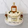 Sofisticado pastel redondo con glaseado crema y un llamativo escurrimiento (drip) dorado metálico. Al frente exhibe una gran etiqueta comestible de Coñac Hennessy Very Special. Está decorado con fresas bañadas en chocolate blanco con el logo de Hennessy, rosetones de crema y perlas doradas. El toque final lo dan una botella real de Hennessy en la corona y dos miniaturas en la base, junto a una placa que dice "Happy Birthday Beautiful". Castillo's Cake Shop.