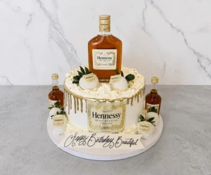 Licor3 Sofisticado pastel redondo con glaseado crema y un llamativo escurrimiento (drip) dorado metálico. Al frente exhibe una gran etiqueta comestible de Coñac Hennessy Very Special. Está decorado con fresas bañadas en chocolate blanco con el logo de Hennessy, rosetones de crema y perlas doradas. El toque final lo dan una botella real de Hennessy en la corona y dos miniaturas en la base, junto a una placa que dice "Happy Birthday Beautiful". Castillo's Cake Shop.
