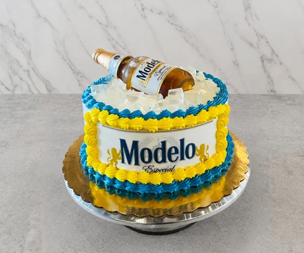 Divertido pastel redondo diseñado para simular una hielera. Los laterales están cubiertos con una etiqueta comestible de Cerveza Modelo Especial, enmarcada por gruesos rosetones entrelazados en colores amarillo y azul intenso. La parte superior está cubierta con cubos de gelatina transparente que imitan hielos, sobre los cuales descansa una botella real de Cerveza Modelo semi-inclinada. Castillo's Cake Shop.