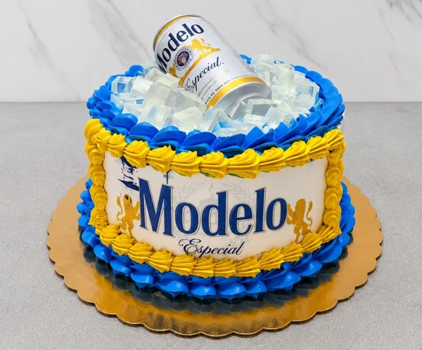 Llamativo pastel redondo que simula una hielera rebosante. Sus costados están cubiertos con el logo clásico de Cerveza Modelo Especial impreso en oblea, rodeado por decorativos rosetones en azul eléctrico y amarillo. La superficie está llena de grandes cubos de gelatina transparente simulando hielo, de donde emerge una lata real de Cerveza Modelo. Castillo's Cake Shop.