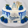 Pastel redondo con un suave diseño en tonos blanco y azul difuminado, destacando un vibrante escurrimiento (drip) azul oscuro. En el frente luce la etiqueta comestible de Tequila Don Julio Blanco. Está decorado con fresas bañadas en una brillante cobertura azul metálica con destellos, rosetones de crema blanca, un letrero dorado de "Happy Birthday" y una botella miniatura real de Don Julio en la corona. Castillo's Cake Shop.