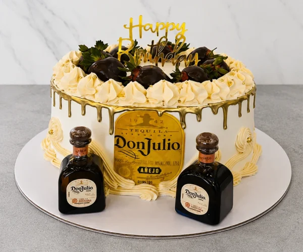Lujoso pastel redondo con cobertura crema suave y un elegante escurrimiento (drip) dorado brillante. Al frente presenta una gran etiqueta comestible de Tequila Don Julio Añejo. La parte superior está adornada con un anillo de rosetones de crema con perlas doradas y fresas frescas bañadas en chocolate oscuro, acompañadas de un letrero dorado de "Happy Birthday". En la base flanquean dos botellas miniatura reales de Tequila Don Julio. Castillo's Cake Shop.