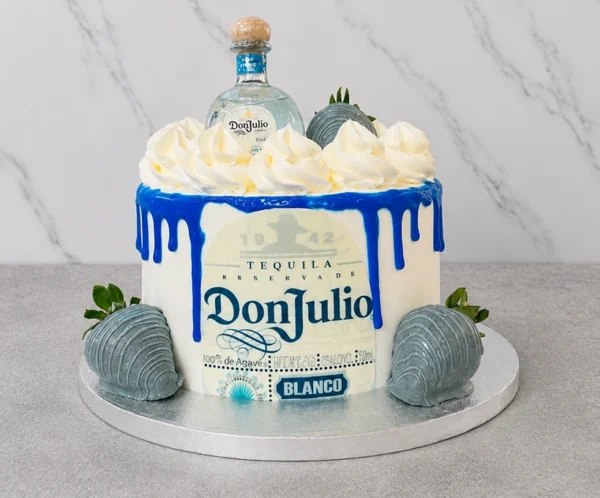 Sofisticado pastel redondo con base blanca y un intenso escurrimiento (drip) azul eléctrico brillante. Al frente exhibe la icónica etiqueta comestible de Tequila Don Julio Blanco. La corona está decorada con grandes copos de crema blanca, y el diseño se complementa con fresas bañadas en una cobertura azul grisáceo perlado. En el centro superior luce una botella miniatura real de Tequila Don Julio Blanco. Castillo's Cake Shop.