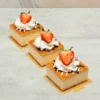 Mini Churro Chesse Cakes. Tres porciones cuadradas individuales de cheesecake servidas sobre pequeñas bases doradas. Tienen una superficie dorada y horneada (estilo churro), finamente decoradas con líneas de chocolate y glaseado blanco, un rosetón de crema batida y coronadas con la mitad de una fresa fresca.