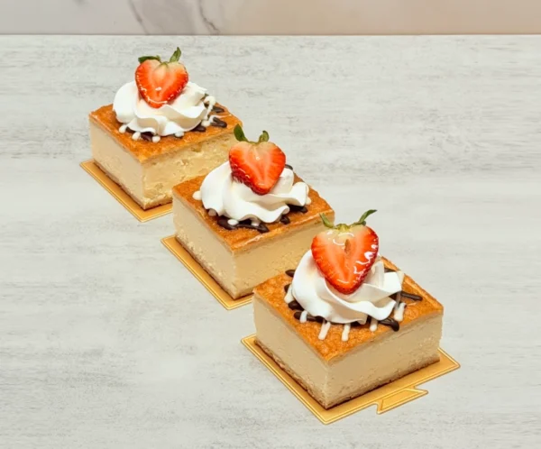 Mini Churro Chesse Cakes. Tres porciones cuadradas individuales de cheesecake servidas sobre pequeñas bases doradas. Tienen una superficie dorada y horneada (estilo churro), finamente decoradas con líneas de chocolate y glaseado blanco, un rosetón de crema batida y coronadas con la mitad de una fresa fresca.