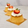 Mini Flanes. Tres clásicos flanes napolitanos individuales presentados sobre bases cuadradas doradas. Lucen una textura porosa característica y una cubierta de caramelo líquido, decorados en la superficie con un copo de crema batida y la mitad de una fresa fresca.