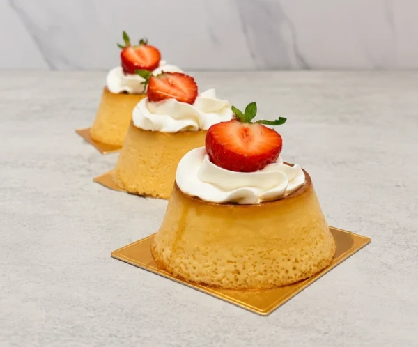 Mini Flanes. Tres clásicos flanes napolitanos individuales presentados sobre bases cuadradas doradas. Lucen una textura porosa característica y una cubierta de caramelo líquido, decorados en la superficie con un copo de crema batida y la mitad de una fresa fresca.