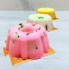 Mini gelatinas. Tres gelatinas individuales con un diseño tipo rosca (bundt cake), presentadas sobre bases cuadradas doradas. Aparecen en tres colores: rosa al frente, blanca al centro y amarilla al fondo. Todas tienen estilo mosaico, dejando ver pequeños cubos de gelatina oscura encapsulados en su interior.