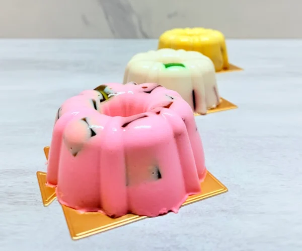 Mini gelatinas. Tres gelatinas individuales con un diseño tipo rosca (bundt cake), presentadas sobre bases cuadradas doradas. Aparecen en tres colores: rosa al frente, blanca al centro y amarilla al fondo. Todas tienen estilo mosaico, dejando ver pequeños cubos de gelatina oscura encapsulados en su interior.