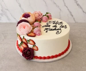 Pastel redondo con cobertura blanca y un borde inferior de glaseado rojo. Está decorado con hermosas flores en tonos rosa y durazno, fresas frescas (algunas de ellas bañadas en chocolate rosa con líneas blancas) y el mensaje "Feliz dia de las Madres" escrito en elegante caligrafía negra. Castillo's Cake Shop.