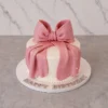 Mujer10 Tierno y elegante pastel redondo con cobertura blanca lisa y un borde inferior de perlas blancas. El gran protagonista del diseño es un enorme moño de fondant rosa con un acabado perlado y efecto de tela drapeada, adornado con diminutas perlas blancas. Sobre la base del pastel se lee el mensaje "HAPPY BIRTHDAY" en letras plateadas. Castillo's Cake Shop.